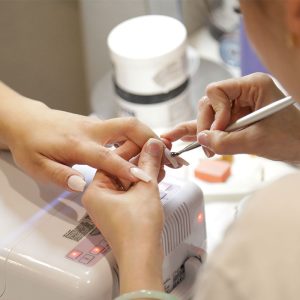 A beauty training specialist applying white nail polish to another person's hand, in a salon environment, as part of a nail technician course.